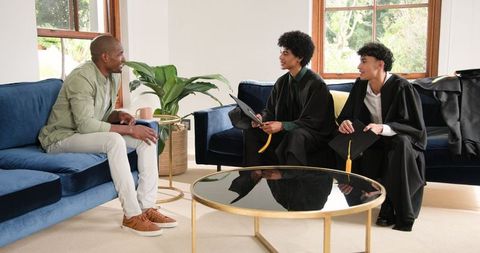 Proud father congratulating sons in graduation gowns at home