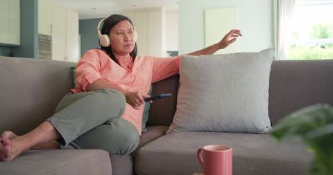 Mature woman relaxing on sofa wearing headphones and holding remote in modern living room