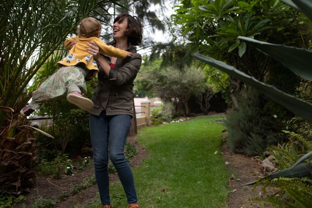 Young mother lifting her baby in a lush garden, both smiling and enjoying the moment. Ideal for use in family-oriented advertisements, parenting blogs, and lifestyle magazines. Perfect for illustrating themes of motherhood, bonding, and outdoor activities.