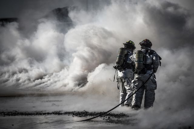 Firefighters are seen in full protective gear working together to manage intense smoke, showcasing the urgency and bravery required in firefighting and emergency response efforts. This image is ideal for promoting awareness about the importance of firefighting, emergency preparedness, and teamwork in hazardous conditions. It can be used for educational purposes, news articles on emergency services, safety training materials, and public service announcements.
