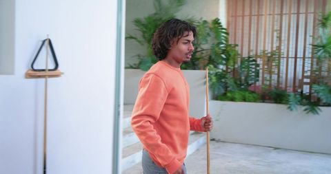 Young man holding wooden pole and walking through modern courtyard wearing orange crewneck