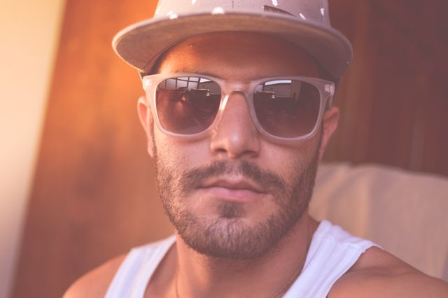 Portrait style featuring young man with beard wearing sunglasses and a hat, showing modern casual fashion trends. Suitable for lifestyle, fashion, and urban culture themes.