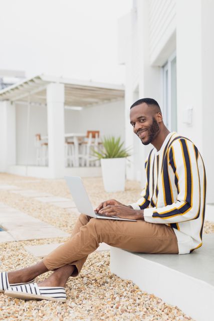 African-American man sitting outdoors by the pool, smiling while using a laptop. He is dressed casually in a striped sweater and brown pants, enjoying a relaxed atmosphere. Ideal for use in content related to remote work, modern lifestyle, leisure activities, technology, and summer relaxation.