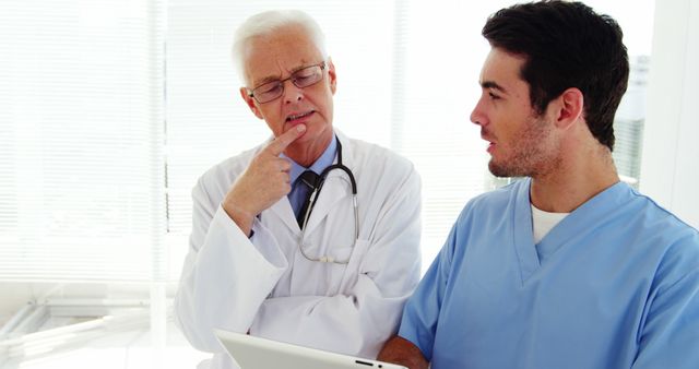 A senior Caucasian male doctor is in deep thought while discussing with a young Caucasian male nurse, with copy space. Their focused interaction suggests a professional healthcare setting where critical medical decisions or patient care strategies are being considered.
