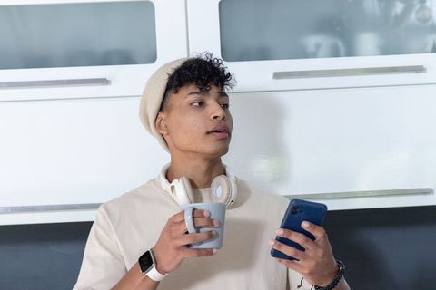 African american man holding smartphone and mug in minimalist kitchen with headphones