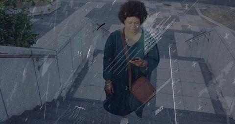 Mid-adult woman climbing urban stairs while checking smartphone, carrying crossbody bag