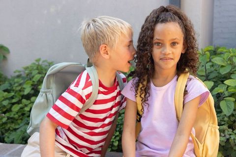 Multiracial children whispering on bench with backpacks, playful school break outdoors