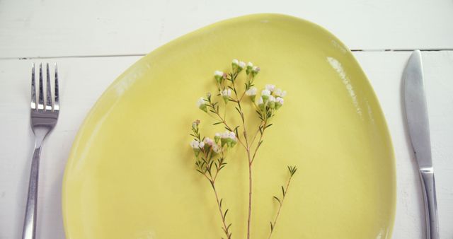 A yellow plate with a delicate floral arrangement is presented on a white wooden surface, with a fork and knife placed on either side, with copy space. The setting suggests a theme of spring or a concept related to fresh, minimalist dining.