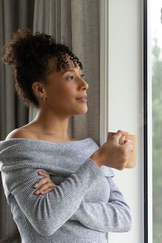 African american woman enjoying morning coffee by window in cozy off-shoulder sweater