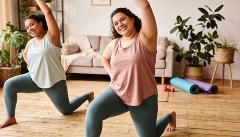 Hispanic women lunging in living room during partner home workout with yoga mats, dumbbells