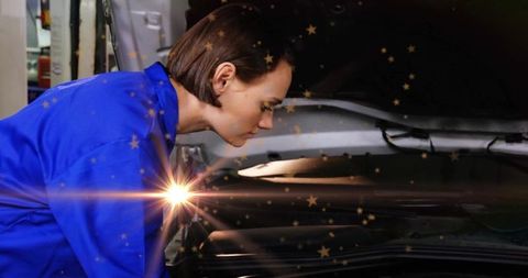 Woman mechanic inspecting engine bay leaning into hood in cobalt coveralls with lens flare
