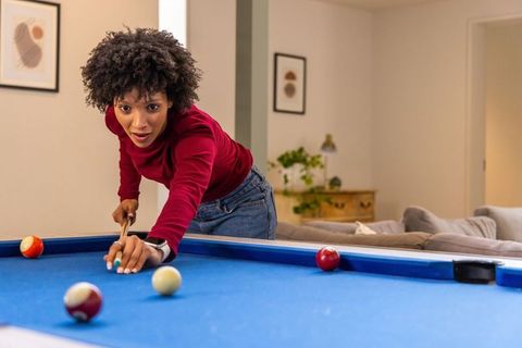 Focused african american woman playing pool leaning over blue table in cozy living room