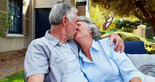 A senior Caucasian couple shares a tender kiss while sitting outside their home, with copy space. Their affectionate moment captures the enduring love and companionship that can flourish in later years.