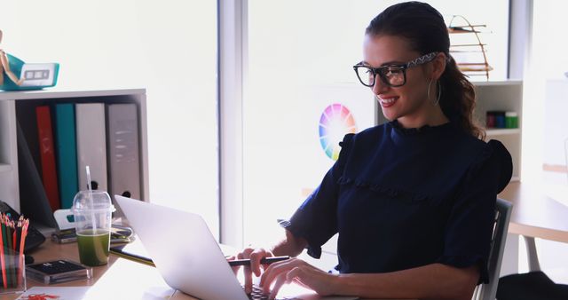 Woman sitting at desk in modern office, typing on laptop and smiling. Bright, sleek workspace with organized shelves and a glass of green smoothie on the table. Ideal for illustrating business, technology, productivity, and contemporary work environments. Useful for articles on remote work, office ergonomics, and work-life balance.