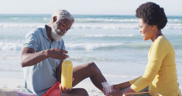 Senior couple sitting on the beach, pouring juice and enjoying the sunny day near the ocean. Elderly man and woman relaxing outdoors, perfect for travel, vacation, lifestyle, and health-related content.