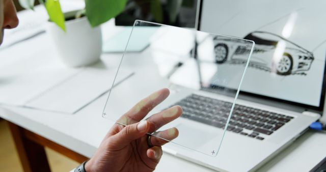 Designer examining transparent glass panel in a modern workspace with a laptop displaying a car design. Useful for themes such as futuristic technology, creative workspaces, product design, and innovation in office environments.