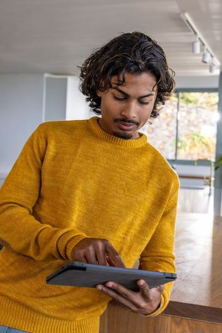 Asian man using tablet in contemporary kitchen wearing mustard sweater working from home