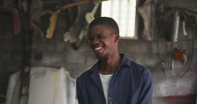 Smiling African American mechanic works in rustic workshop, surrounded by tools and auto parts. He wears blue coveralls and exudes a positive, welcoming vibe. This image can be perfect for showcasing tradesmen, workshops, automotive repair services, and community stories spotlighting hardworking individuals.