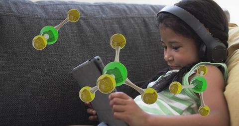 Young girl using tablet with headphones exploring augmented reality molecules on couch