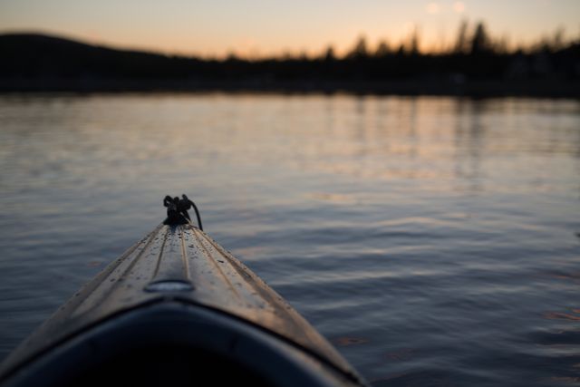 Kayak prow cutting through still water during a serene sunset, creating a silhouette effect with gentle ripples. Used for promoting outdoor activities, nature retreats, advertisements on adventure getaways, or blog posts about peaceful travel experiences.