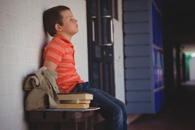 Sad boy with eyes closed sitting on bench by wall in corridor at school