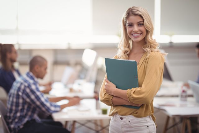 Young businesswoman smiling and holding a file in a modern, creative office environment. Ideal for use in business, corporate, and professional contexts, showcasing teamwork, confidence, and a positive work atmosphere.