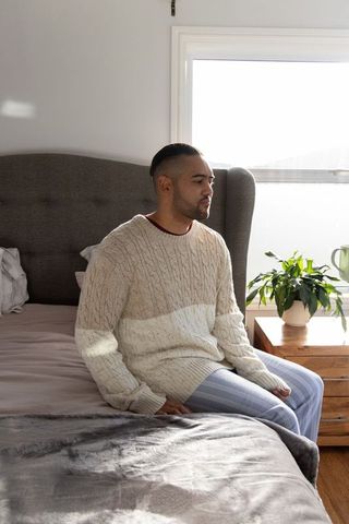 Man sitting on bed facing window wearing cream sweater and lounge pants in serene bedroom