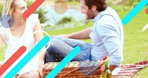 Couple enjoying lakeside picnic, chatting on blanket with wicker basket and wine bottle