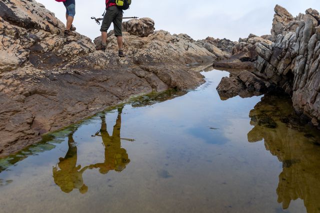 Senior couple hiking on rocky terrain, holding hands and carrying backpacks with hiking poles. Their reflections are visible in a water pool on the rocks. Ideal for use in travel blogs, adventure magazines, fitness promotions, and retirement lifestyle content.