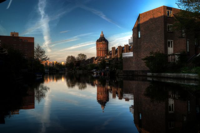 A peaceful scene of an urban waterway at sunset where a historic water tower and residential buildings reflect in the canal water. The soft, dim sky with streaky clouds adds to the tranquility of the scene. Useful for travel brochures, urban landscape collections, architectural presentations, and calm, reflective themed content.
