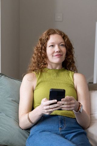Redhead woman sitting on couch scrolling smartphone wearing green top and jeans