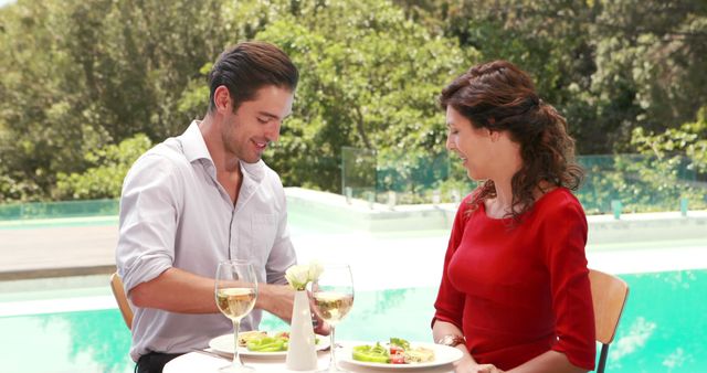 A young Caucasian couple enjoys a romantic lunch by a poolside, with copy space. Their cheerful engagement over a meal in a serene outdoor setting suggests a moment of leisure and connection.