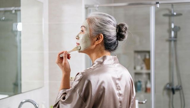 Senior asian woman applying green facial mask while wearing silk robe in modern bathroom