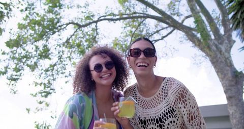 Laughing african american women enjoying refreshing summer drinks in backyard party with sunglasses
