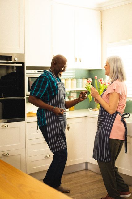 Senior couple wearing aprons dancing in kitchen, creating a joyful and loving atmosphere. Perfect for articles or ads related to retirement living, family life, senior activities, and lifestyle content. Great for use in brochures, websites, or social media posts promoting happiness and togetherness in older age.