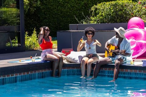 African american friends relaxing by pool playing guitar and sipping drinks on sunny summer day