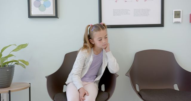 A young girl with pigtails sitting in a clinic waiting area, displaying a thoughtful expression. She is wearing a light colored jacket and pants and is seated on a modern chair. Medical posters and a thermostat are visible on the wall behind her. This image can be used to depict themes related to healthcare, pediatric care, waiting, anticipation, mood, and children's healthcare services.