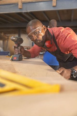 African american carpenter leaning over workbench holding cordless drill in workshop