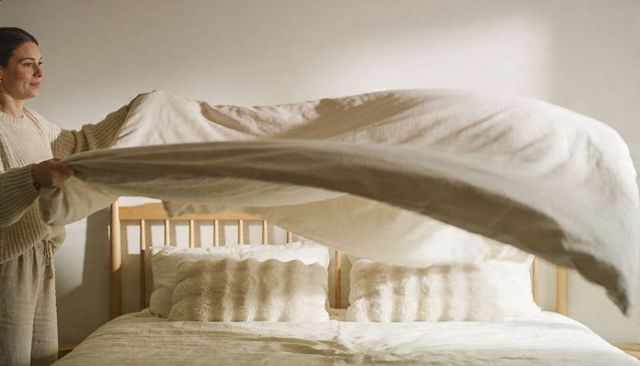 Woman Making Bed with Linen Duvet in Sunlit Minimalist Bedroom Featuring Slatted Wood Headboard