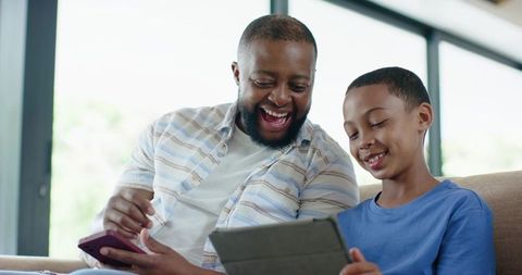 African american father and son sharing tablet and laughing together on sofa