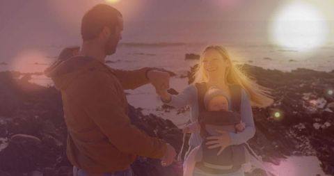 Family walking on rocky shore at sunset with baby in front carrier and golden lens flare