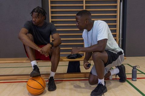 African american coach coaching male basketball player resting on gym bench with stopwatch