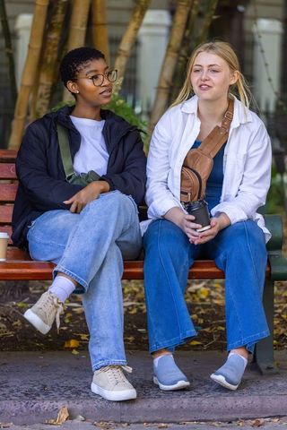 Two friends sitting on park bench chatting and drinking coffee with crossbody bags