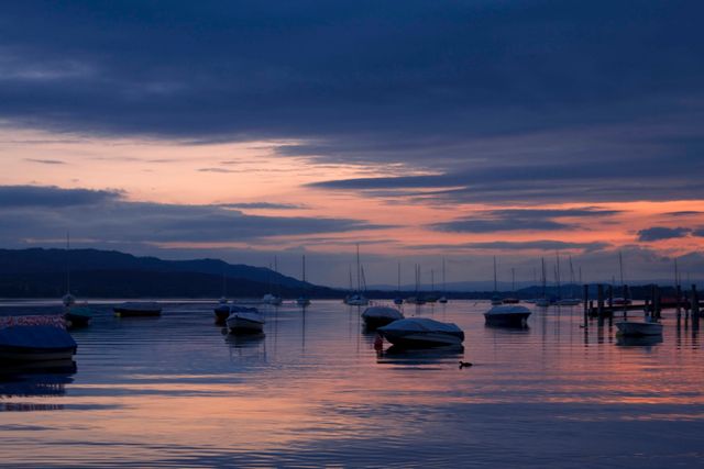 Tranquil sunset scene with moored boats casting reflections on calm harbor water. Docks and distant mountains are visible under an idyllic twilight sky. Ideal for themes involving relaxation, nature, scenic beauty, and travel destinations.
