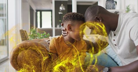 Father and son sharing tender forehead moment on sofa, cupping head, golden particle swirl