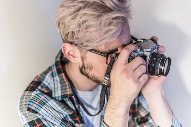 A young man is using a vintage film camera to take pictures in a studio setting. He is wearing a plaid shirt and glasses, focusing on his shot. This image is ideal for representing creative pursuits, hobbies, and the revival of traditional photography techniques. Use it for blogs, ads, or articles about photography enthusiasts, artistic expression, and professional photographers.
