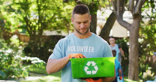 Man collecting recyclable materials in green bin, wearing gloves and volunteer shirt. The sunny outdoor setting with trees adds vibrant greenery, promoting eco-friendly initiatives. Ideal for content promoting environmental conservation, volunteering, community clean-up events, or sustainability projects.