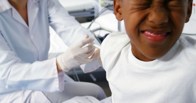 Young boy receiving a vaccine injection from pediatric doctor in a clinic setting. The healthcare professional is wearing a white coat and gloves, and the child is wincing during the procedure. Suitable for use in medical websites, vaccination campaigns, educational materials on immunization, and promoting pediatric healthcare services.