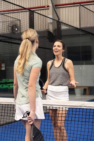Two women chatting at net on indoor paddle court, exchanging friendly tips after match