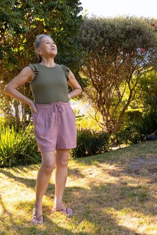 Senior asian woman standing in sunlit backyard garden wearing olive top, pink shorts